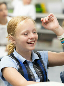 girl in school uniform sits at a desk and raises her hand while smiling