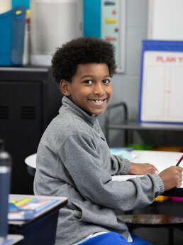 student sitting at desk in gray fleece holding a pencil  and smiling