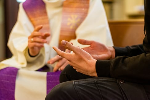 close up showing hands of a man sitting in confession with a priest in the background making a sign of absolution